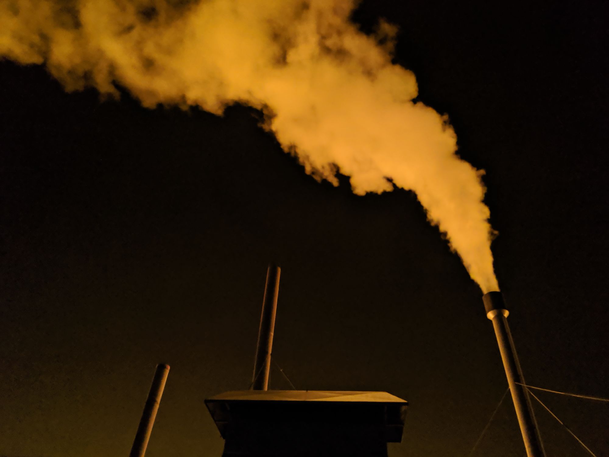 Photo of a trail of smoke coming from a chimney, mostly consisting of water vapor, coming from burned natural gas to warm a greenhouse. You can see the orange-ish glow of the greenhouse fluorescent lights reflecting on the smoke.